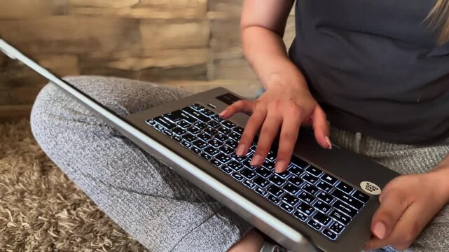 Young businesswoman sitting comfortably on the floor at home, typing on a laptop with a backlit keyboard, engaged in online work, freelance projects, or remote education studies