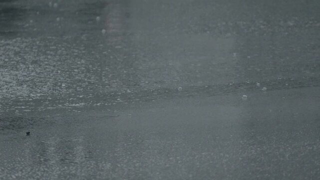 Rain falls steadily on a wet surface creating ripples and reflections during a cloudy day