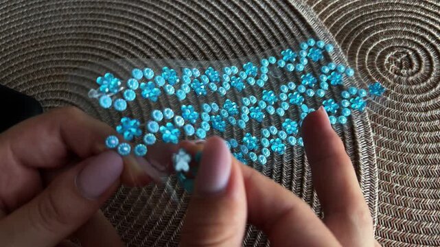 Woman's hands meticulously arranging a decorative floral pattern of adhesive blue rhinestones and pearls on a transparent strip, preparing for a creative craft project