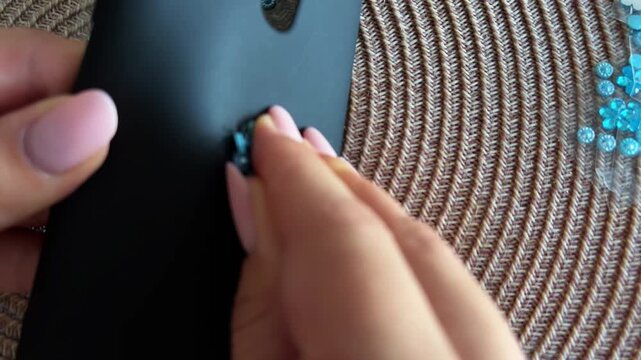 Woman's hands removing blue rhinestone flower sticker from a black smartphone case revealing scratches and damage, showcasing product wear and tear concept