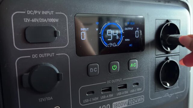 Portable power station being operated by a woman's hand, showing the digital display with battery level, input, and output wattage, while connecting a plug to the ac outlet