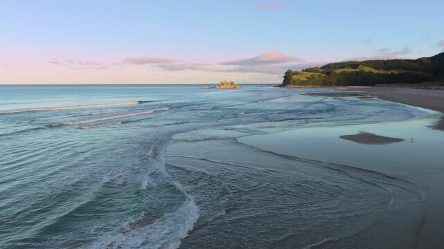 Waves wash ashore at Opoutere Beach, New Zealand, showing a serene coastal landscape with green hills under a peaceful sunset.
