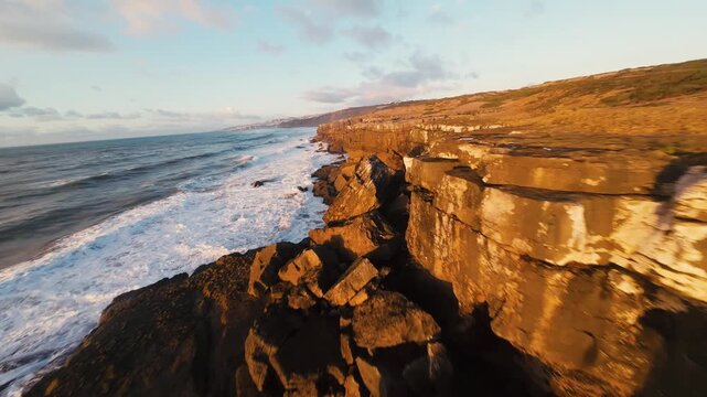 Rugged cliffs meet crashing waves under golden dusk light. Sea foam dances against weathered stones, echoing nature's power. Sunlight glows warmly over ancient rock, painting a serene coastal scene
