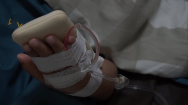 Close-up photo of a patient's hand resting on a padded splint in a hospital bed. The hand has an intravenous (IV) catheter inserted and secured with medical bandages and tape. 