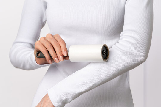 Close up of woman hands removing lint from white clothing with adhesive sticky roller on light background. Concept sustainable clothing upkeep, mindful wardrobe maintenance habits. Copy space, banner