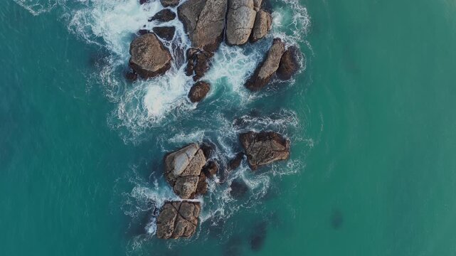 Aerial drone view of turquoise sea waves crashing against rocky coastal boulders, creating white foam in the ocean waters.