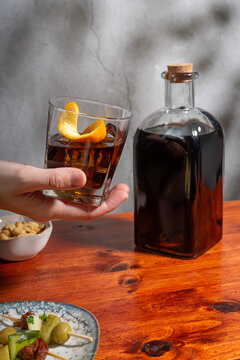 Hand holding a glass of vermouth on the rocks above a wooden table surrounded where there is a bottle, two gildas and a small bowl of peanuts