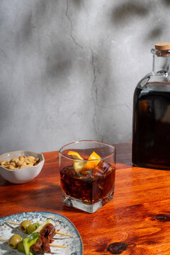 Glass of vermouth on the rocks on a wooden table surrounded by a bottle, two gildas and a small bowl of peanuts