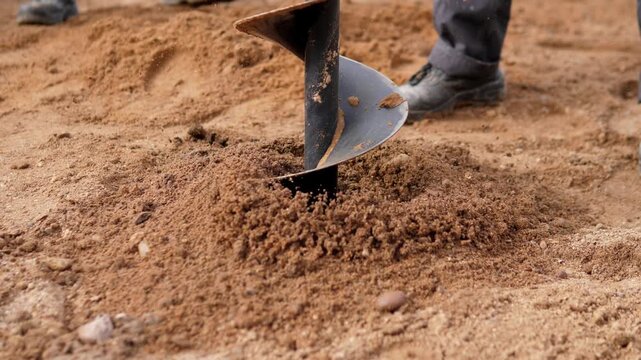 Dark auger bit drives into dry sandy soil, pushing up loose earth while workers boots and trouser legs stand nearby at outdoor construction site, with auger, drilling, soil, sand detail