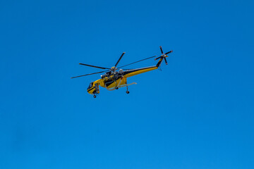 Fototapeta premium Yellow firefighting helicopter with water snorkel flying under blue sky on wildfire mission