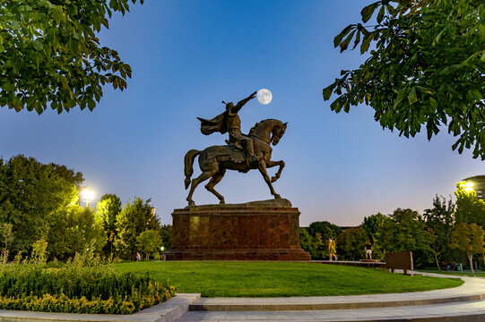 View of the equestrian Statue of Amir Temur set against a clear evening sky with the moon visible above, surrounded by lush park greenery in Shahrisabz, Qashqadaryo Region, Uzbekistan.