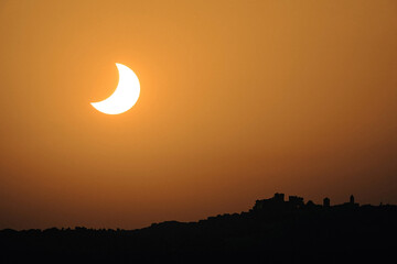 Partial solar eclipse creating a crescent sun against an orange sky, with a dark silhouette of a village and hills below © Manuel