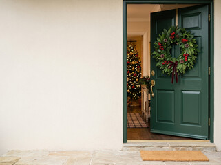Fototapeta premium Open green front door decorated with a holiday wreath revealing a Christmas tree inside. Exterior home entrance with blank light wall providing copy space on the left