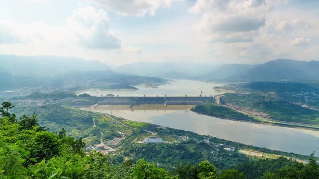 A scenic high angle view shows the massive Three Gorges Dam spanning across the Yangtze River in China with lush green mountains in the background