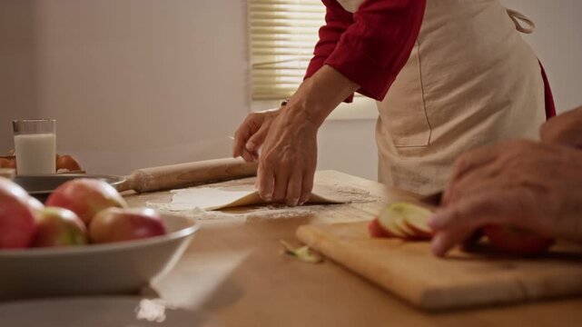 Caucasian senior woman rolling dough with rolling pin on table while senior man cutting fresh apples. Footage works for culinary advertising, bakery commercials, and family lifestyle campaigns