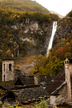 View of the Foroglio waterfall cascading down a rocky cliff behind traditional stone houses with slate roofs in the autumn landscape of Cevio, Ticino, Switzerland.