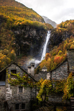 View of Foroglio waterfall cascading behind traditional stone houses with autumn foliage in Cevio, Ticino, Switzerland.