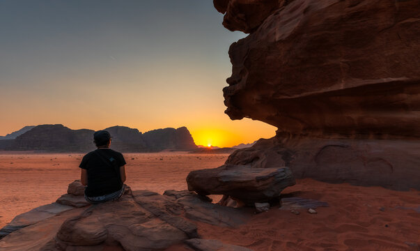 View of a person sitting on a rock formation overlooking the vast desert landscape of the Wadi Rum Protected Area during a vibrant sunset in Wadi Rum Village, Aqaba Governorate, Jordan.