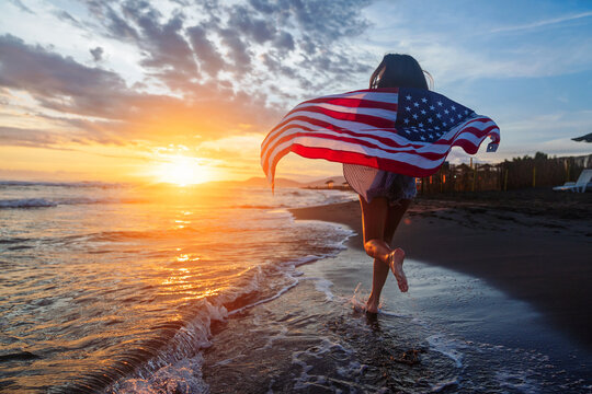 Beautiful patriotic young woman with the American flag held in her standing in the summer sunset