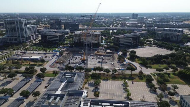 Towering construction cranes dominate open building grounds beside HALL Park within DFW metroplex. Rooftop photovoltaic arrays border multi tier garages in commercial districts, Frisco, Texas