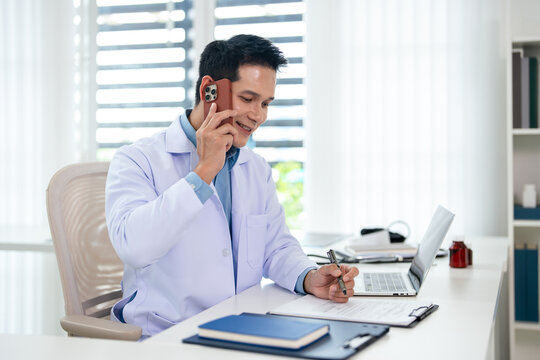 Doctor talking on phone while writing notes in a hospital office surrounded by medical equipment and documents.