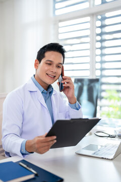 asian male doctor  reads a medical report on a clipboard while talking on the phone in a hospital  office. TeleHealth
