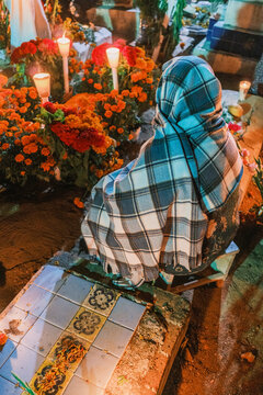 View of a person in a plaid shawl sitting by a grave decorated with orange marigolds and candles during the Day of the Dead celebration in Santa Cruz Xoxocotlan, Oaxaca, Mexico.