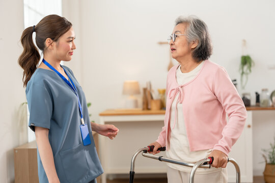 Senior Asian woman practices standing with walker beside young Asian nurse during rehabilitation, supporting mobility, independence and safe active aging inside assisted living senior care center.