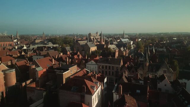 Morning aerial panorama over Bruges, Belgium, displays layered red rooftops, historic houses and distant church towers across the medieval city under clear sky.