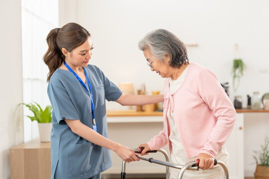 Young Asian nurse supports senior Asian woman using walker during rehabilitation practice, guiding safe mobility, balance and growing independence inside modern assisted living senior care center.