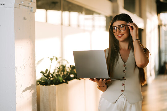 Professional woman holding a laptop while adjusting her glasses in a sunlit office workspace