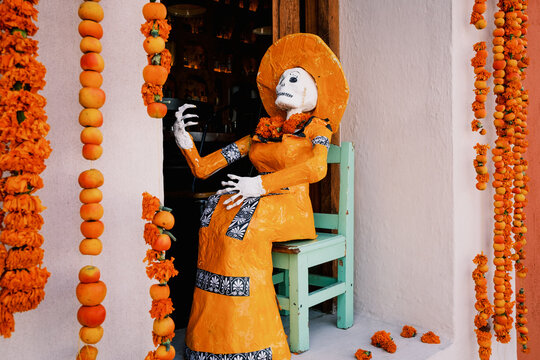 View of a traditional La Catrina figure dressed in orange sitting on a light green chair, surrounded by vibrant marigold garlands during Dia de los Muertos in Oaxaca, Oaxaca, Mexico.