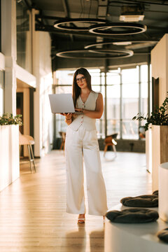 Young woman in a stylish white outfit working on a laptop while standing in a bright modern office