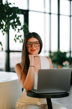 Professional woman in stylish glasses sitting at a table with her laptop in a bright office space