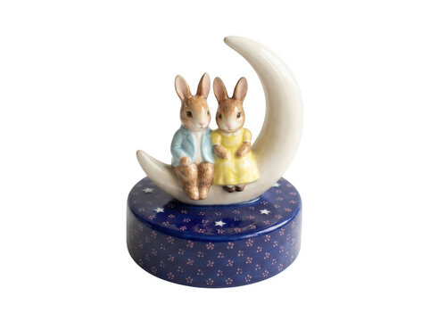 A ceramic music box featuring two cute rabbits sitting on a crescent moon, set on a dark blue base decorated with small white stars and flowers isolated on a transparent background
