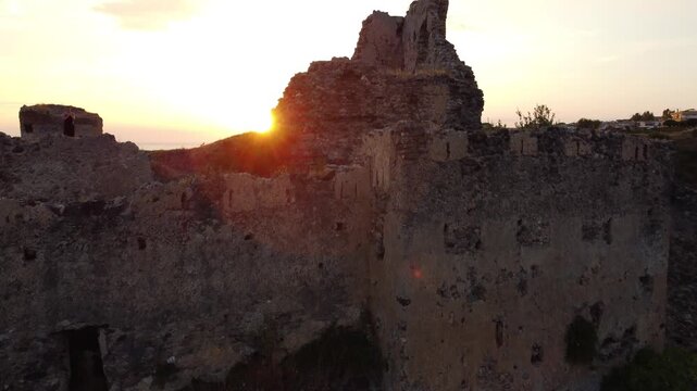 Cinematic aerial reveal shot ascending from ancient castle ruins to a stunning sunset over the Mediterranean Sea and coastal town of Scalea, Calabria, Italy.