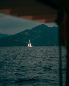 View of a white sailboat sailing on the dark blue sea with the mountainous Gili Trawangan island in the background under a cloudy sky, Pemenang, West Nusa Tenggara, Indonesia.