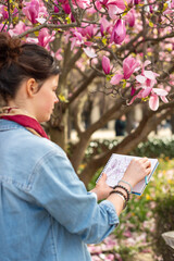 A young female artist sketches in a notebook under a blooming magnolia tree in Paris. The woman draws outdoors in front of pink spring blossoms in a vertical view. © dsbedarieva