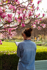 A female artist sketches in a notebook under a blooming magnolia tree in Paris. The back view shows the woman holding a pen and notebook in a vertical frame. © dsbedarieva
