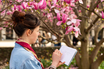 A young female artist sketches in a notebook under a blooming magnolia tree in Paris. The woman holds a pen while drawing outdoors near pink flowers in a horizontal view. © dsbedarieva