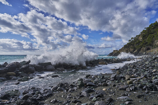 View of waves crashing against a stone breakwater on a rocky beach with a tunnel entrance in the cliffside under a cloudy sky in Liguria, Italy.
