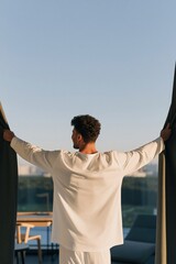 Obraz premium Rear view man opening curtains on a balcony at sunrise with urban skyline for interior background and morning routine banner