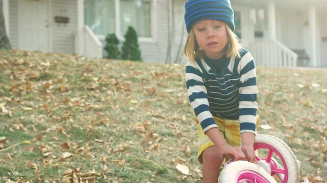 Boy pushing a pink tricycle across a leaf-covered lawn in front of a white house