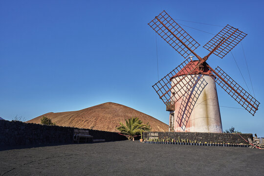 View of a traditional windmill with wooden blades and a red roof set against a volcanic hill and black volcanic soil in Lanzarote, Canarias, Spain.
