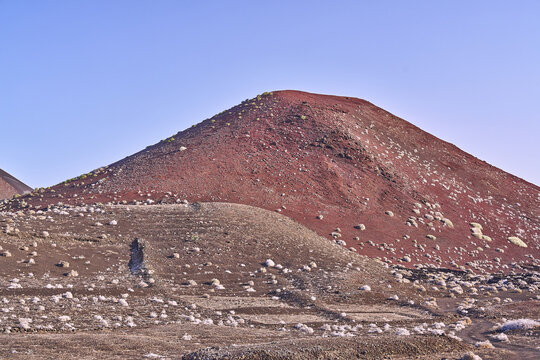 View of a red volcanic cone rising above the rocky terrain and sparse vegetation of the arid landscape in Lanzarote, Canarias, Spain.
