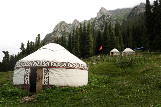 View of Altyn-Arashan yurt set in a lush green mountain meadow with spruce trees and rocky peaks in the background in Altynarasan, Issyk-Kul Region, Kyrgyzstan.