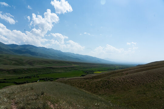 View of the Kilemche valley with rolling green hills, vast grassy plains, and distant mountains under a bright blue sky in Kyzart, Naryn Region, Kyrgyzstan.