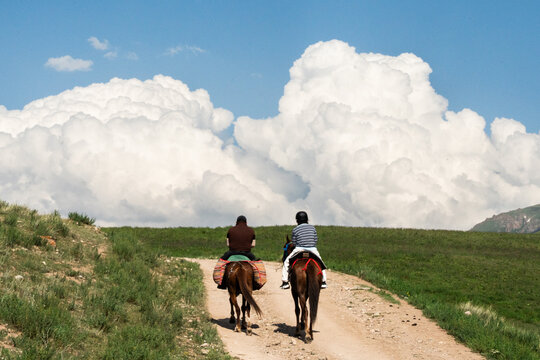 View of two people riding horses on a dirt path through the Kilemche valley, with lush green hills and large cumulus clouds in the sky, Kyzart, Naryn Region, Kyrgyzstan.