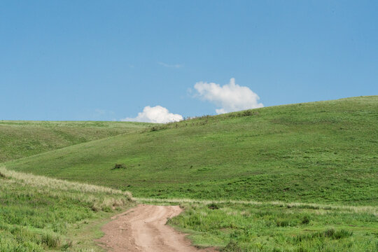 View of a dirt road winding through the rolling green hills of the Kilemche valley under a clear blue sky in Kyzart, Naryn Region, Kyrgyzstan.