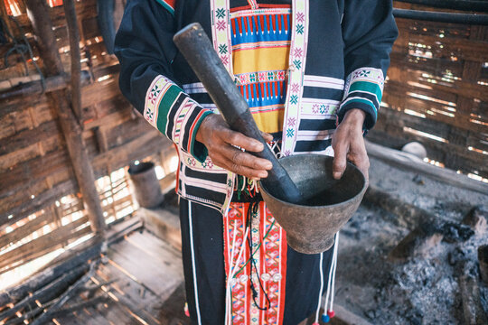 View of a Hmong tribe member wearing traditional embroidered clothing while using a wooden mortar and pestle inside a rustic home in Laos.